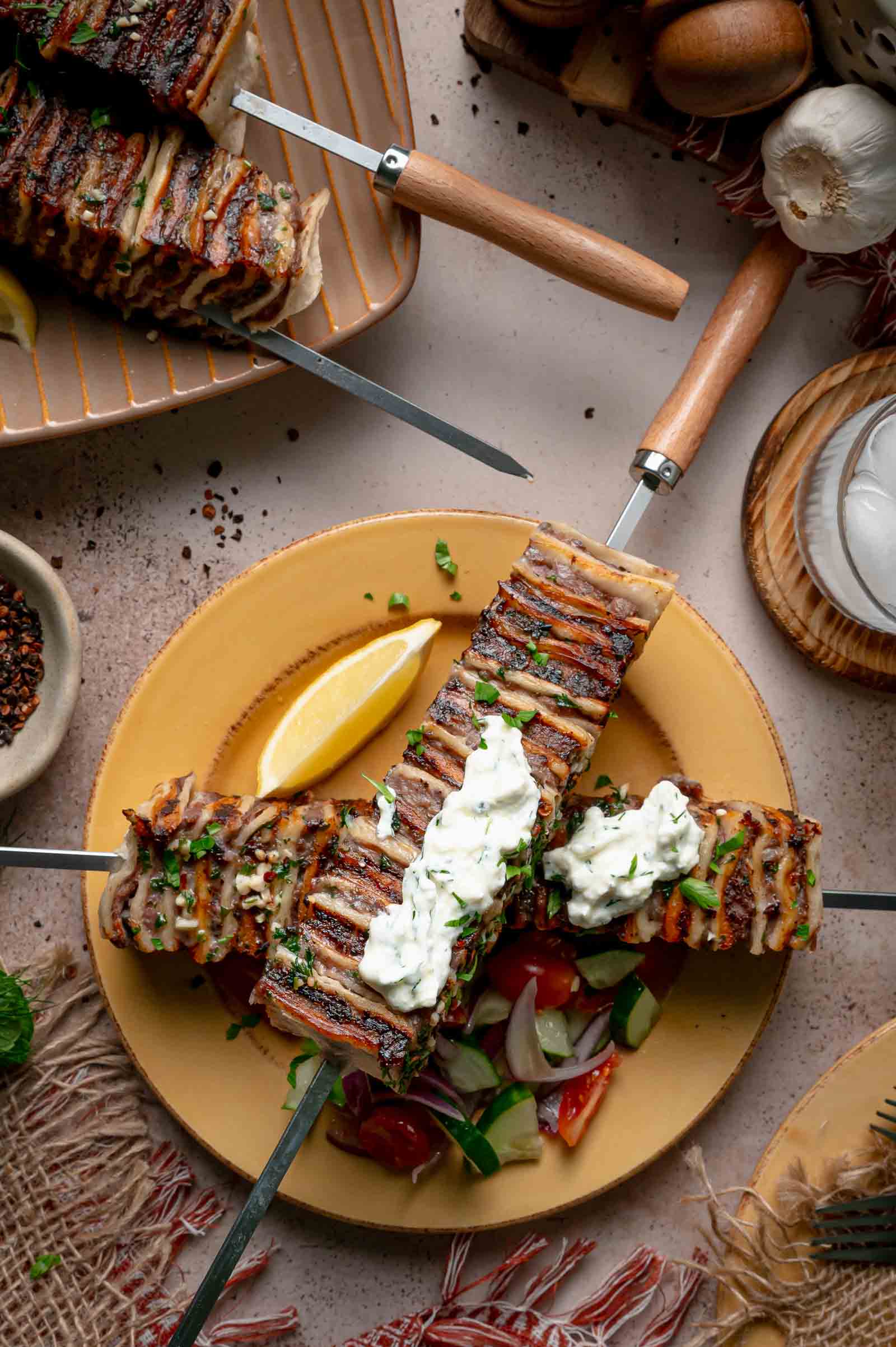Two tortilla kebab skewers crossed on a plate with dollops of haydari, a lemon wedge and chopped cucumber tomato salad.