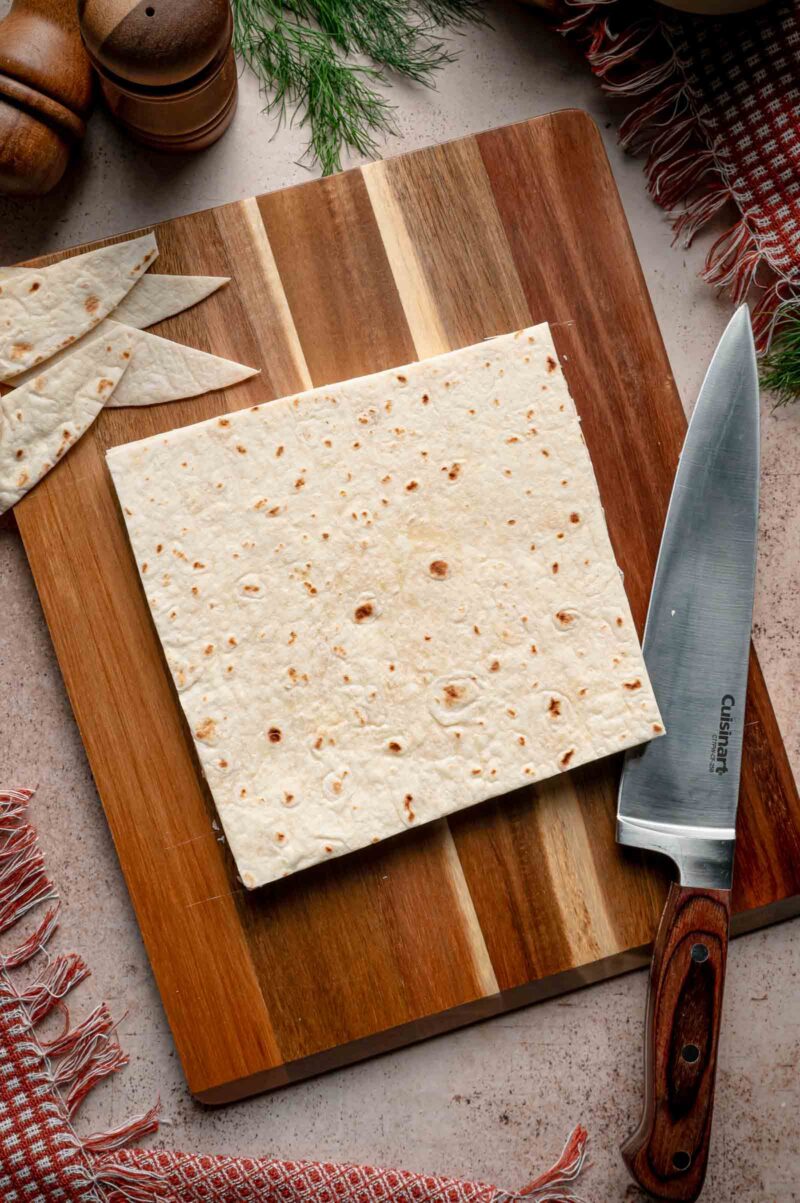 A flour tortilla trimmed into a square with a chef's knife, the rounded offcuts set aside on the board.