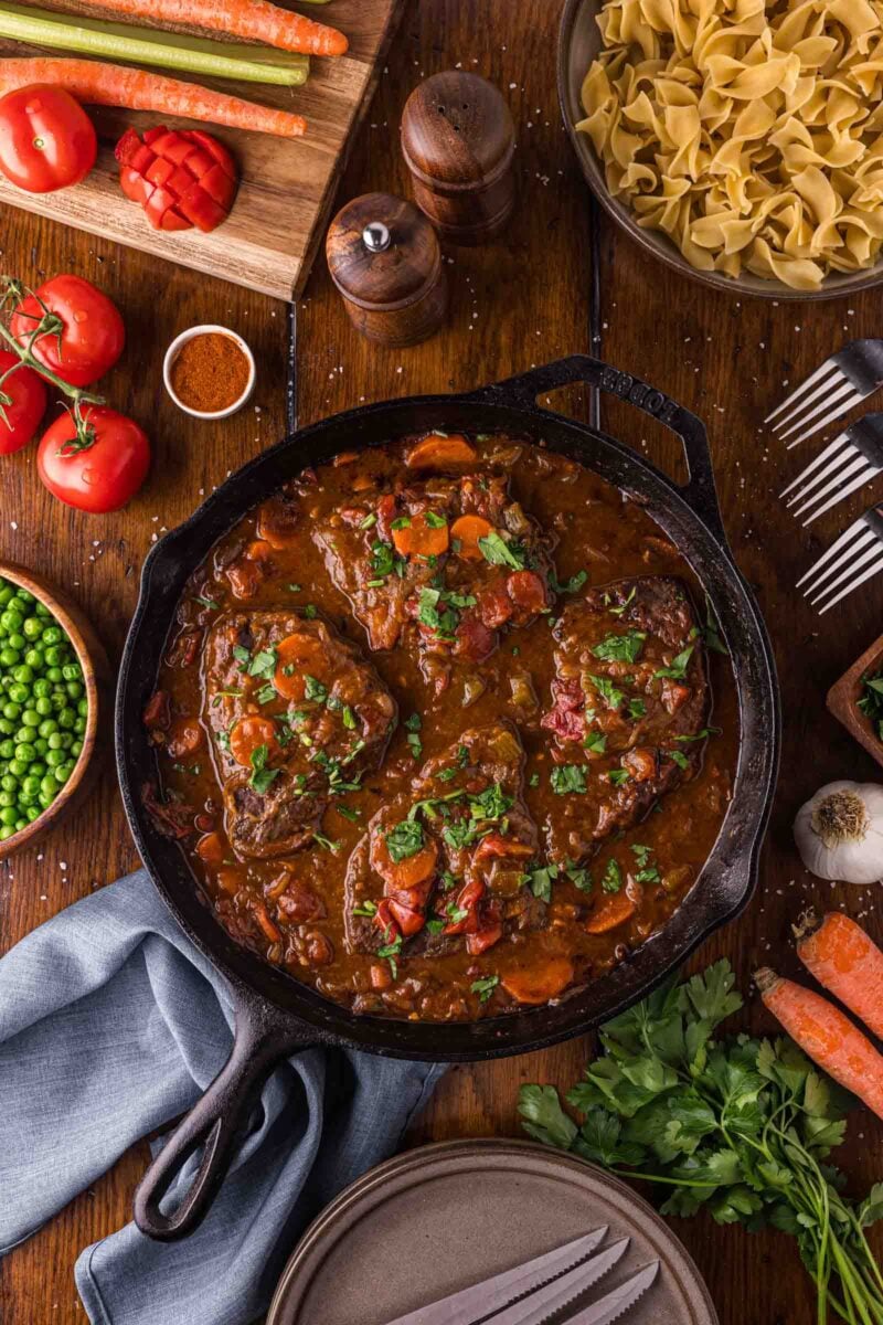 The finished Swiss steak in its cast iron skillet, surrounded by carrots, tomatoes, peas, egg noodles and fresh parsley.
