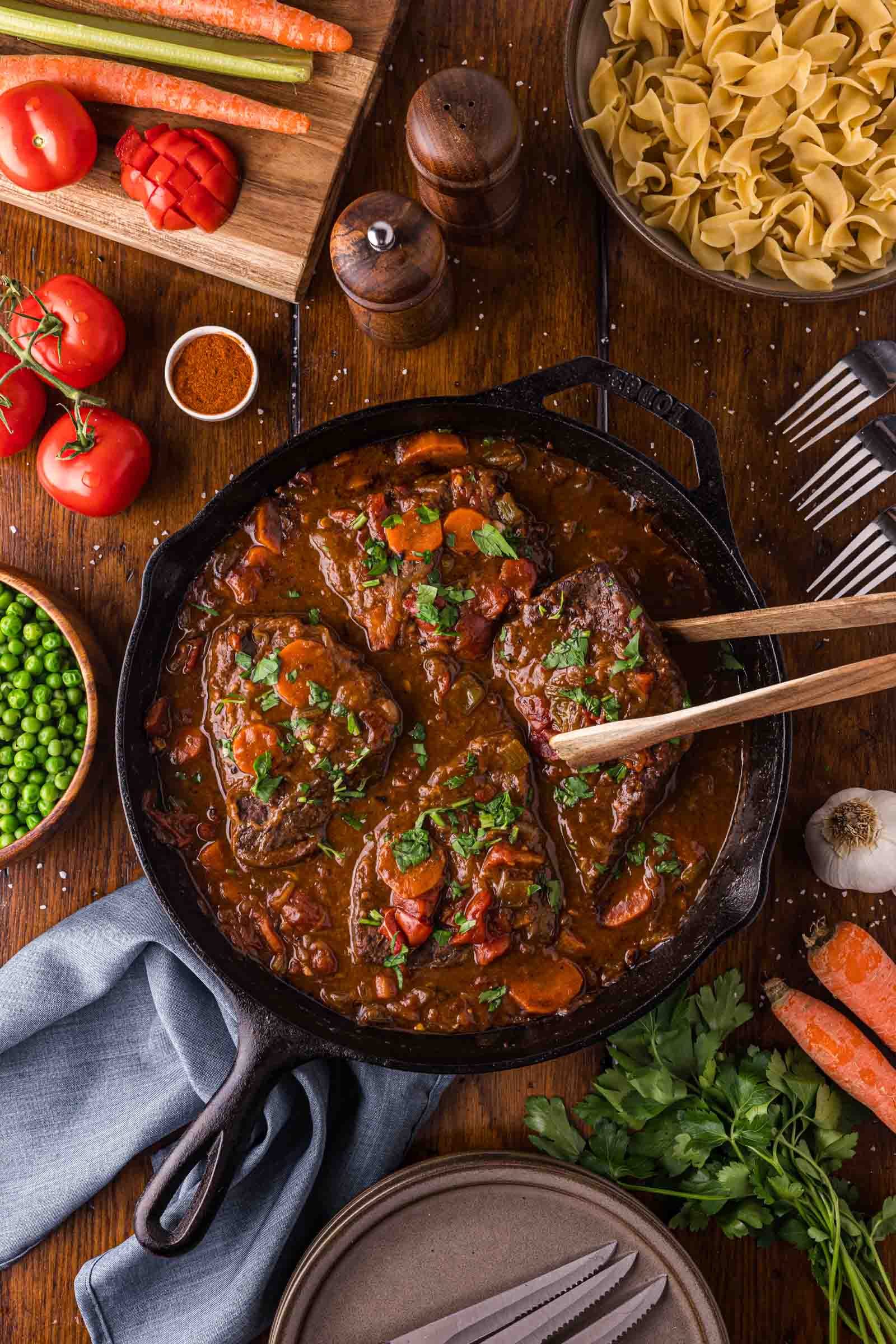 Swiss steak resting in tomato gravy in the skillet with wooden spoons, egg noodles and green peas waiting alongside.