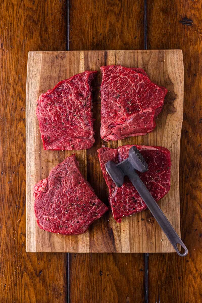 Four seasoned round steak portions laid out on a wooden cutting board with a meat mallet, ready to be pounded even.