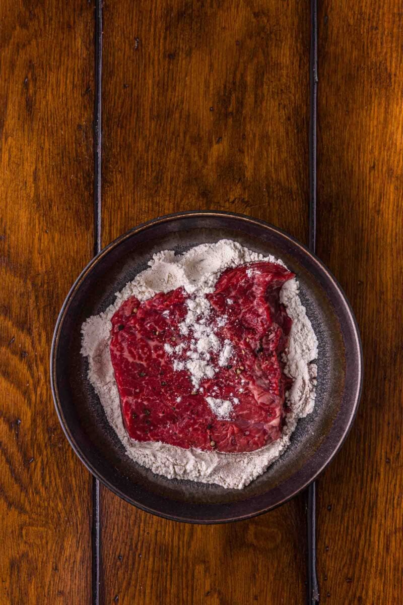 A round steak dredged in seasoned flour in a shallow bowl, coated on both sides before it goes into the pan.