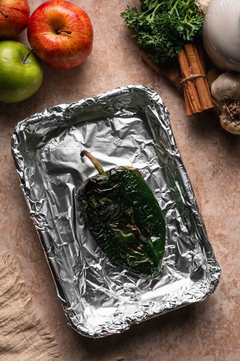 A whole poblano roasted until the skin blisters and blackens, on a foil-lined pan beside apples, parsley and cinnamon sticks.