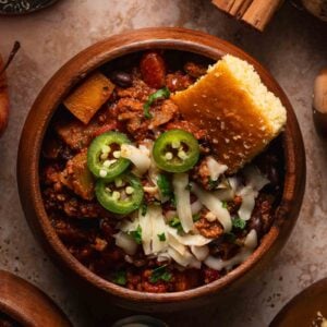A wooden bowl of turkey apple chili with shredded cheese, sliced jalapeno, parsley and a wedge of cornbread.