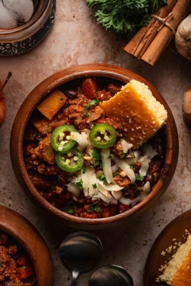 A wooden bowl of turkey apple chili with shredded cheese, sliced jalapeno, parsley and a wedge of cornbread.