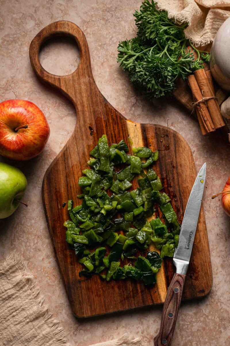 Chopped roasted poblano on a wooden board with a paring knife, apples and cinnamon sticks alongside.