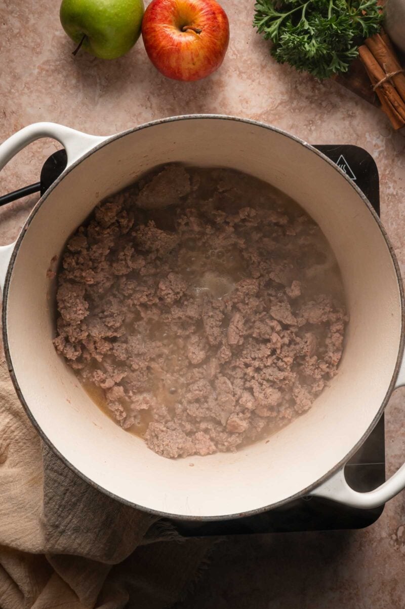 Ground turkey browning in a white Dutch oven, red and green apples and cinnamon sticks on the counter nearby.