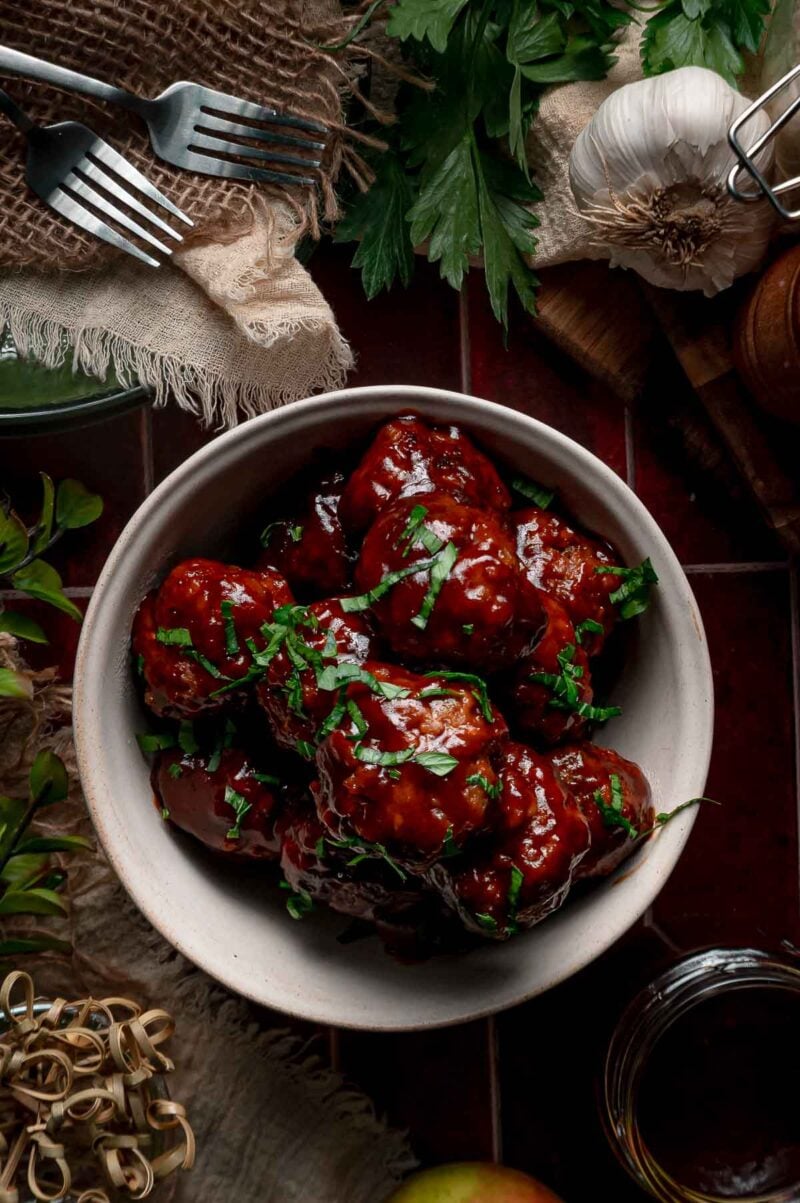 A bowl of apple butter BBQ meatballs topped with chopped parsley, with forks, fresh parsley, garlic and an apple on the table.