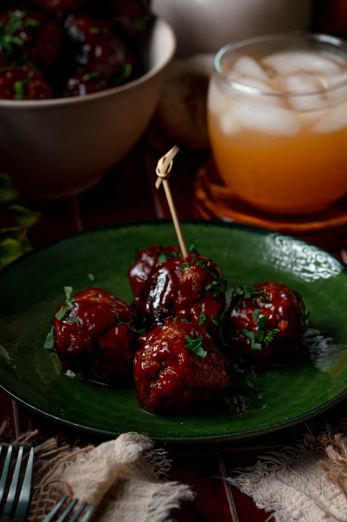 Apple butter BBQ meatballs on a green plate with a knotted bamboo pick, next to an iced drink and the serving bowl.