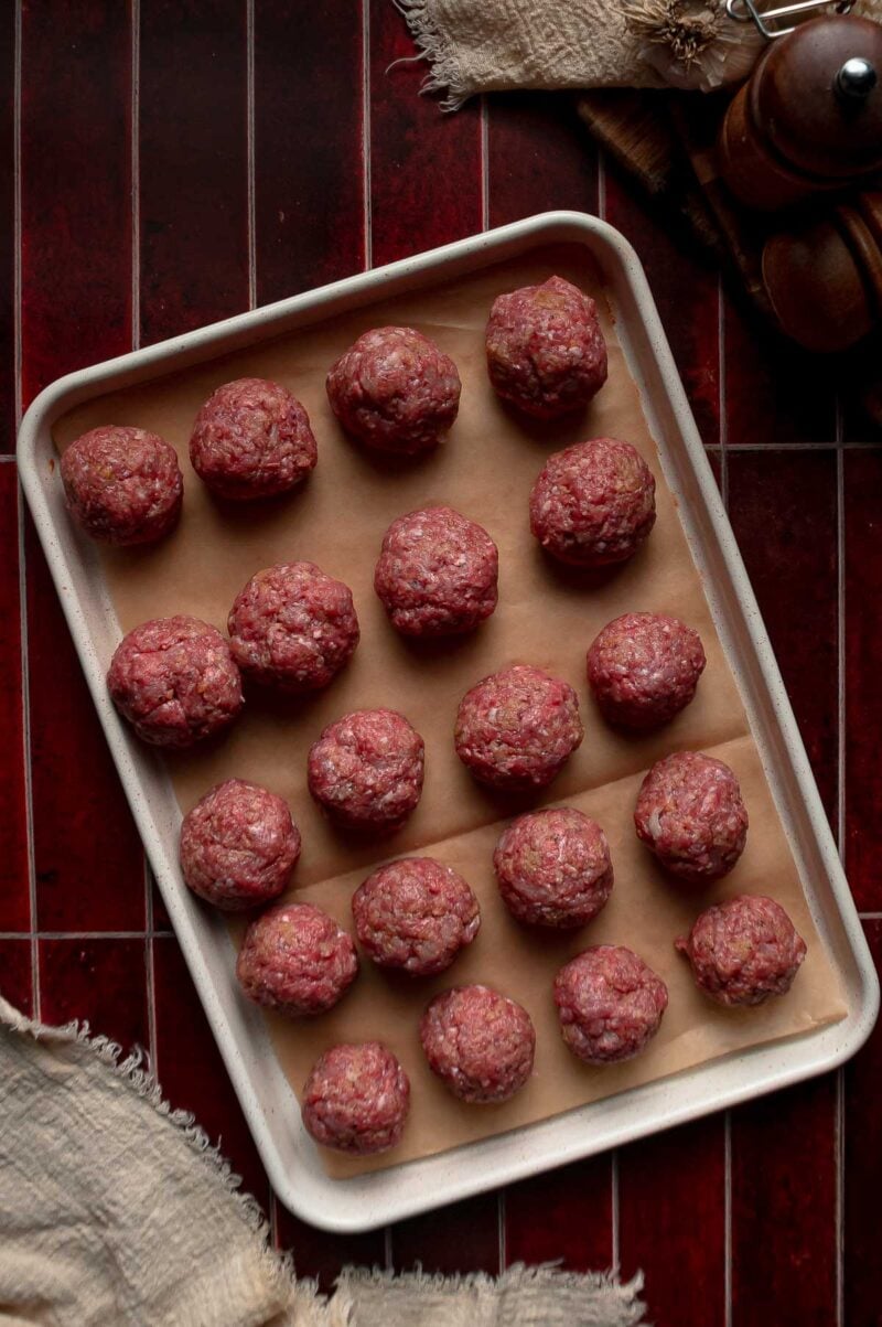 Raw beef and pork meatballs rolled to the same size and lined up on a parchment-lined sheet pan.