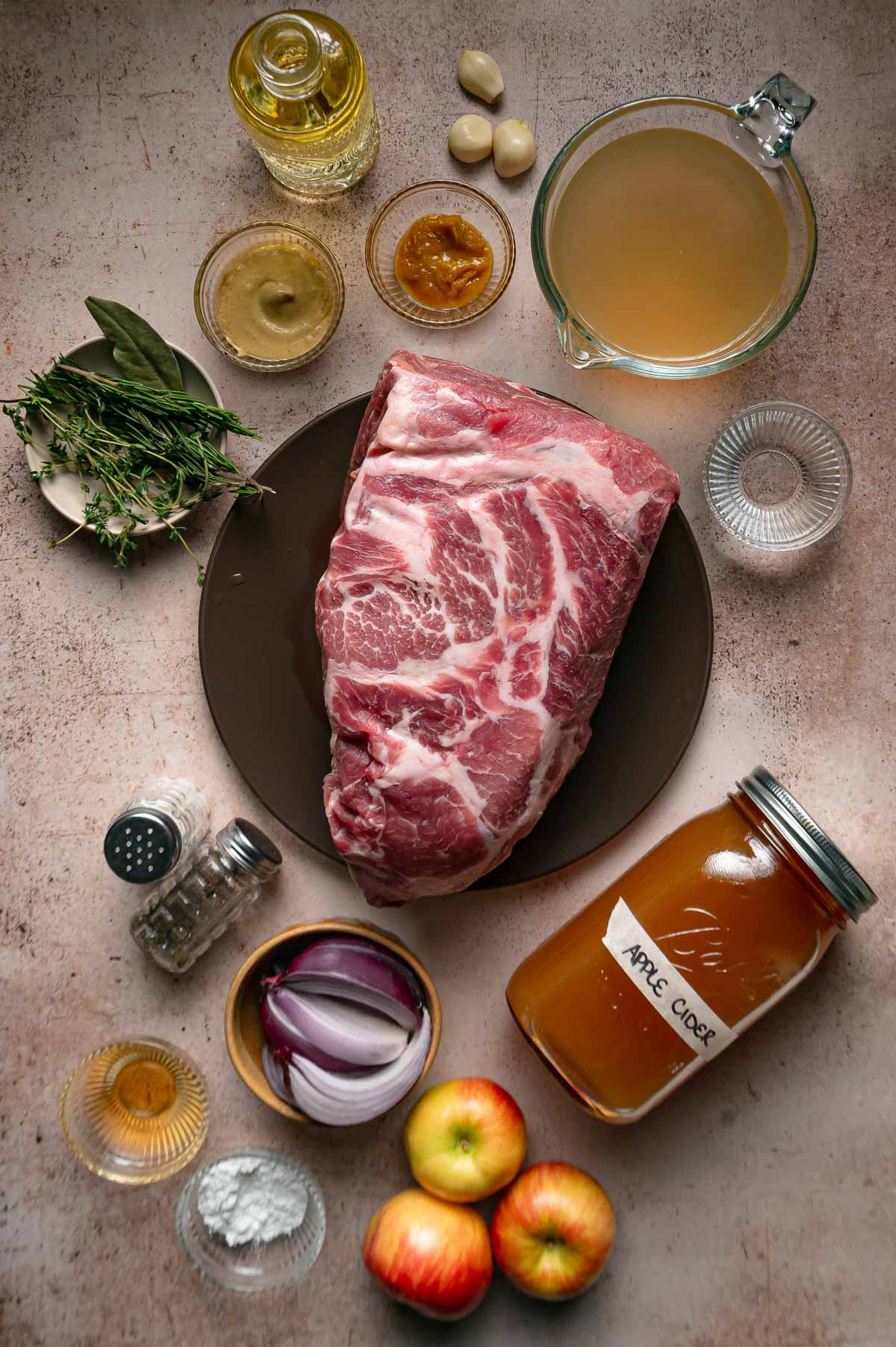 Ingredients for apple cider braised pork: a raw pork shoulder, a jar of apple cider, red apples, red onion, garlic, fresh herbs, broth and seasonings.