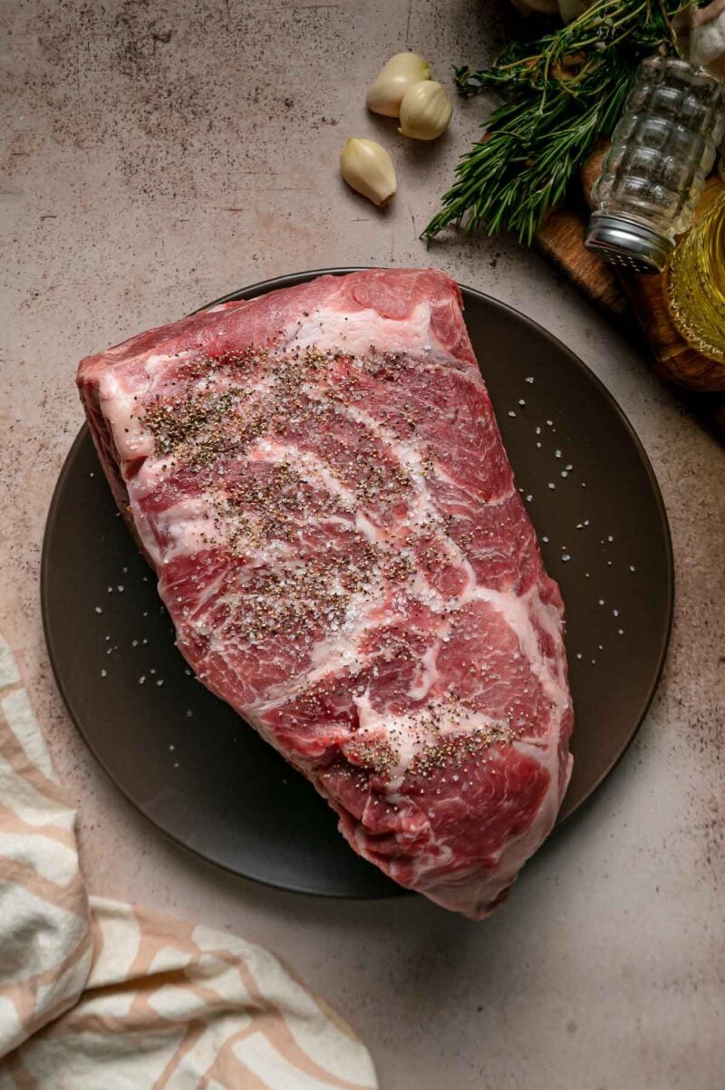 A raw pork shoulder seasoned with salt and pepper on a dark plate, with garlic cloves and herbs nearby.