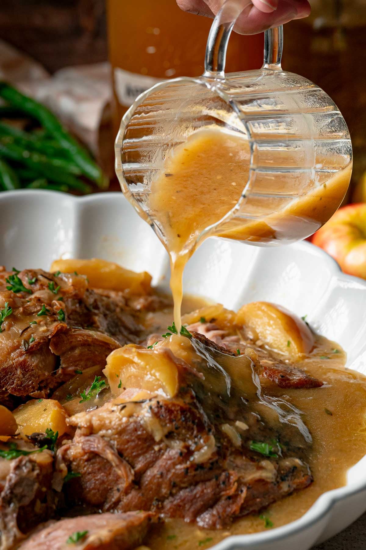 Gravy being poured from a glass pitcher onto a serving of cooked pork and apple slices.