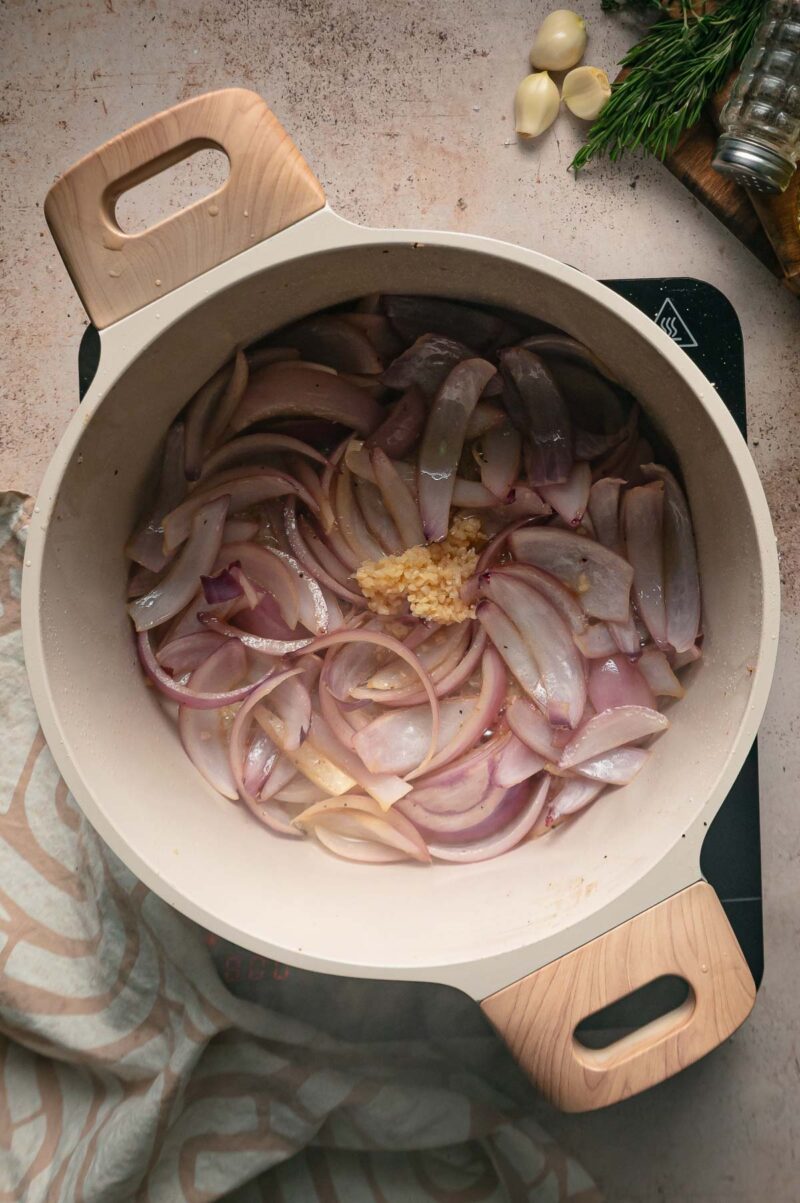 Sliced red onions and minced garlic cooking in a light-colored pot on a stovetop.