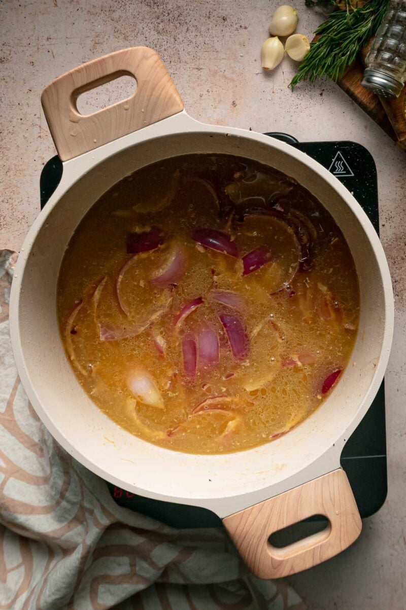A light-colored pot on a burner containing a simmering broth with sliced red onions.