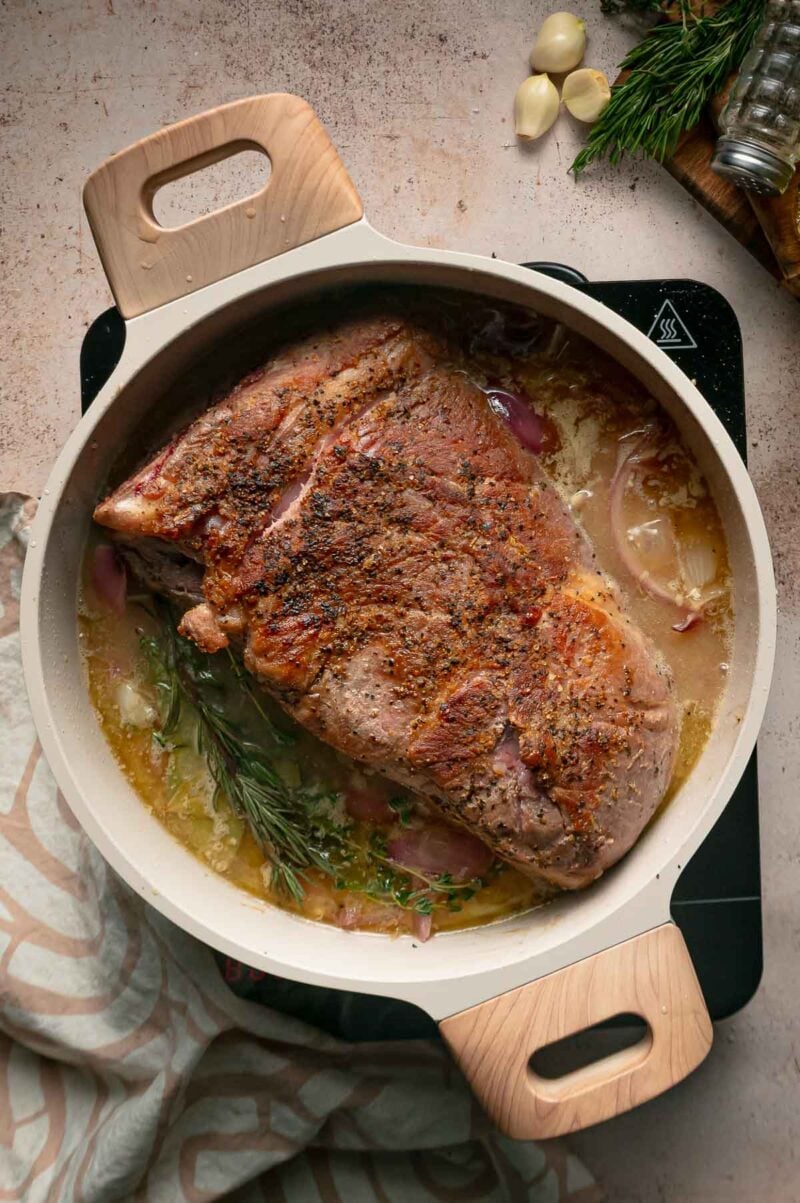 The seared pork shoulder returned to the pot of braising liquid and onions, with fresh herb sprigs tucked alongside.