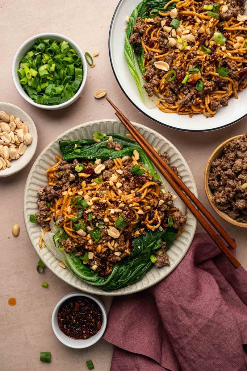Two bowls of ground lamb dan dan noodles surrounded by bowls of green onions, peanuts, chili oil and extra browned lamb.