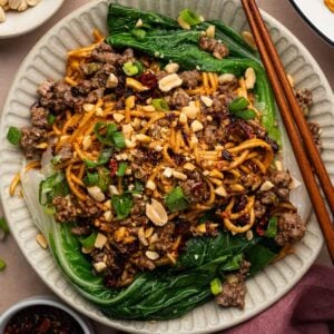 Overhead of ground lamb dan dan noodles on bok choy leaves, scattered with chopped peanuts, green onions and chili flakes.