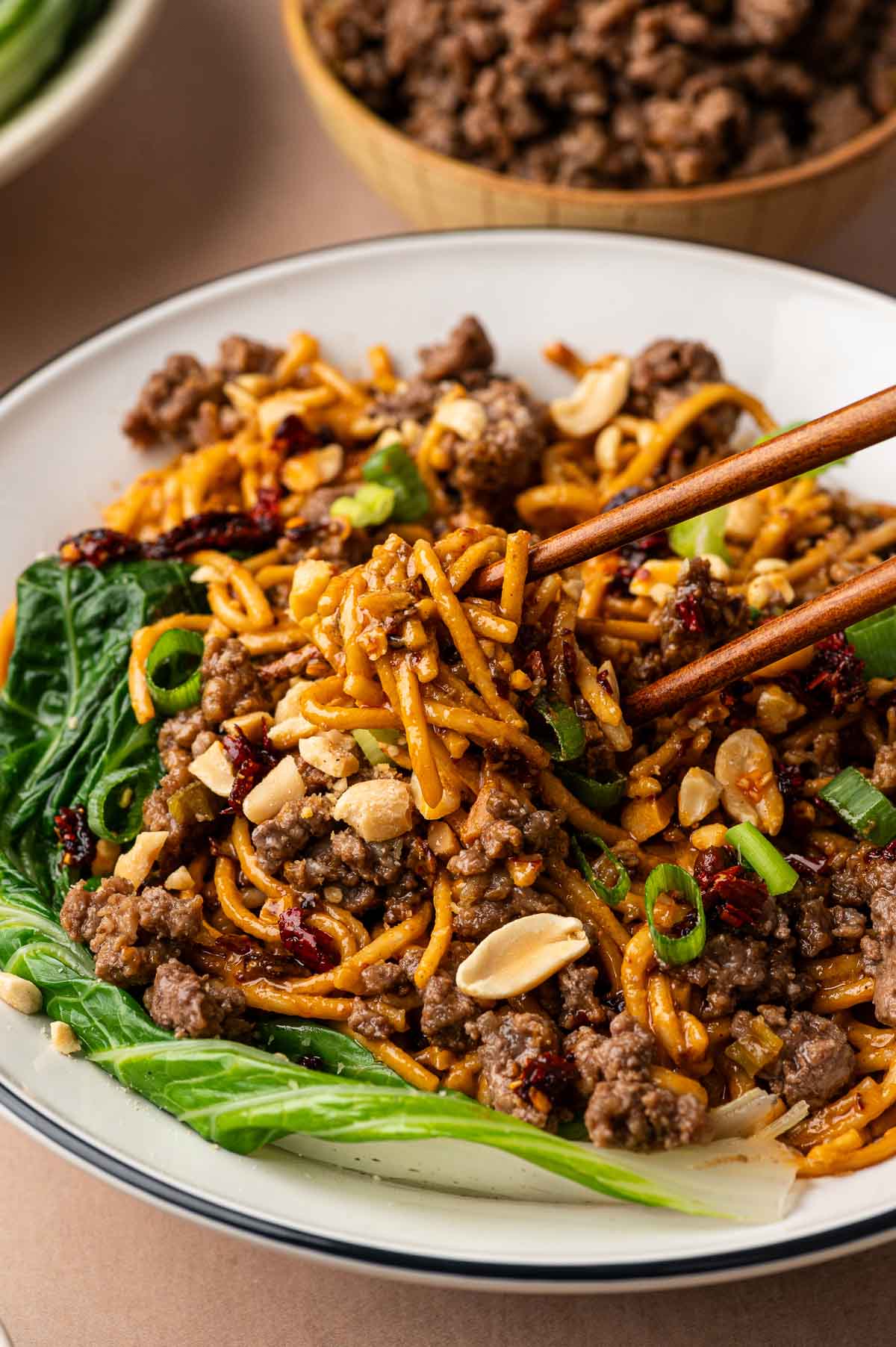 Chopsticks lifting ground lamb dan dan noodles from a bowl lined with bok choy, a dish of browned lamb behind it.
