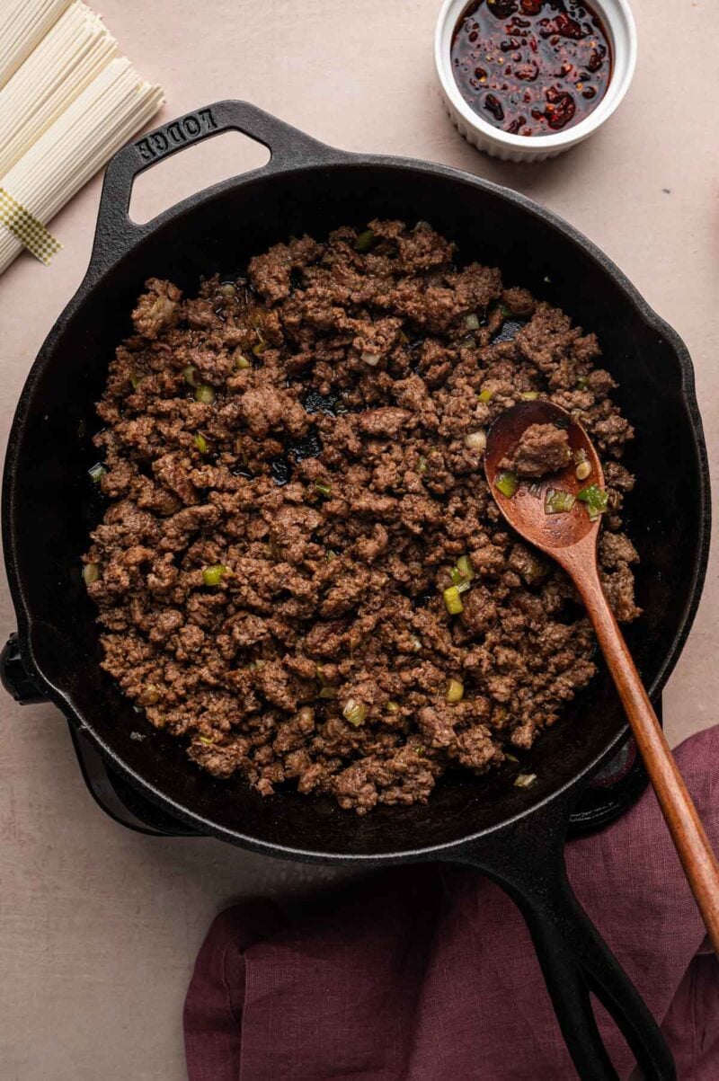 Browned ground lamb with sliced green onion whites in a cast iron skillet, a wooden spoon resting in the pan.