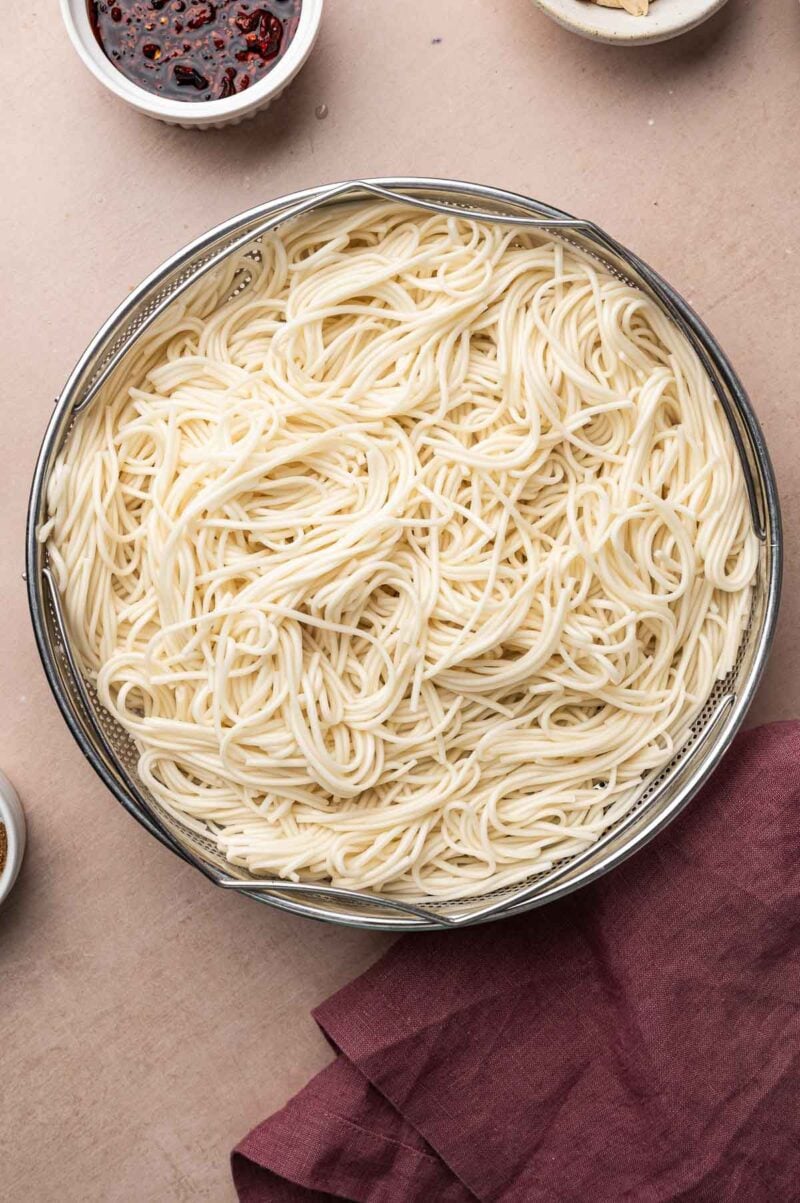 Cooked thin white noodles draining in a metal strainer basket.