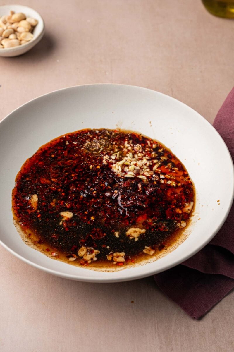 Chili oil, minced garlic and ground Sichuan pepper in a wide white bowl, ready to be whisked into dan dan sauce.