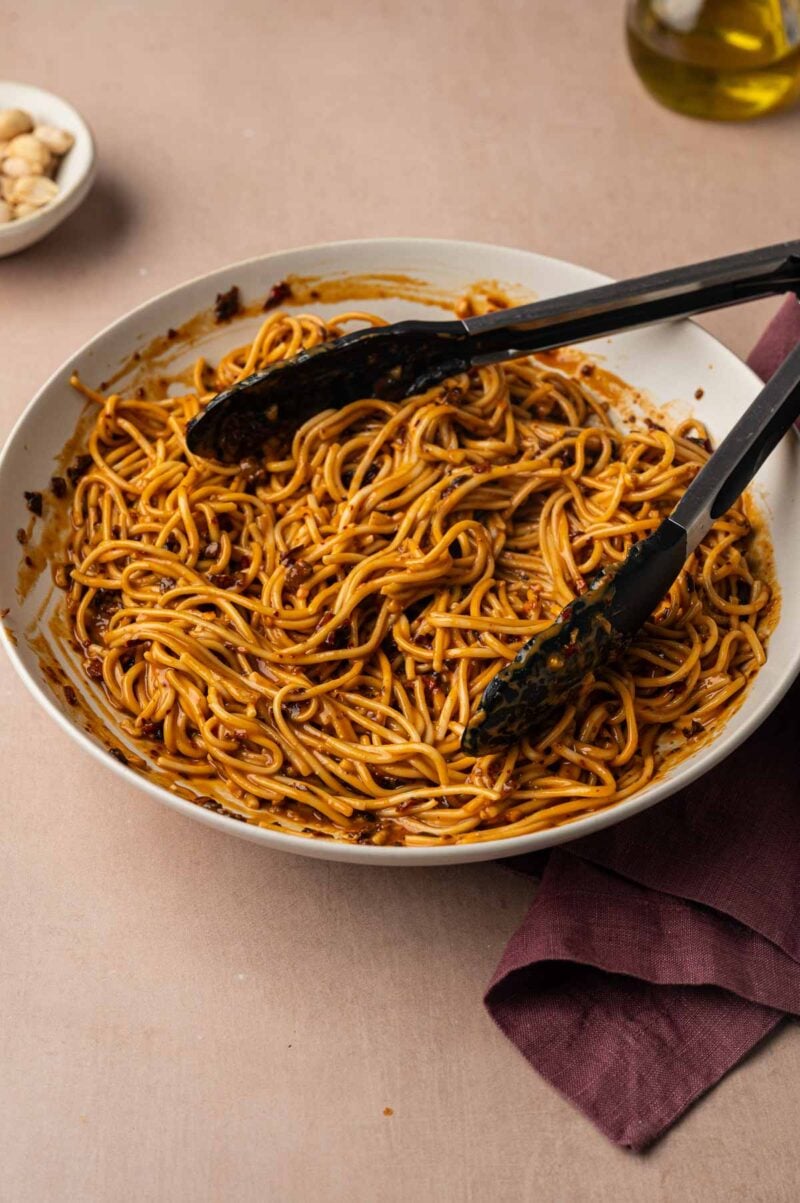 Noodles tossed in dan dan sauce in a white bowl, lifted with black tongs.