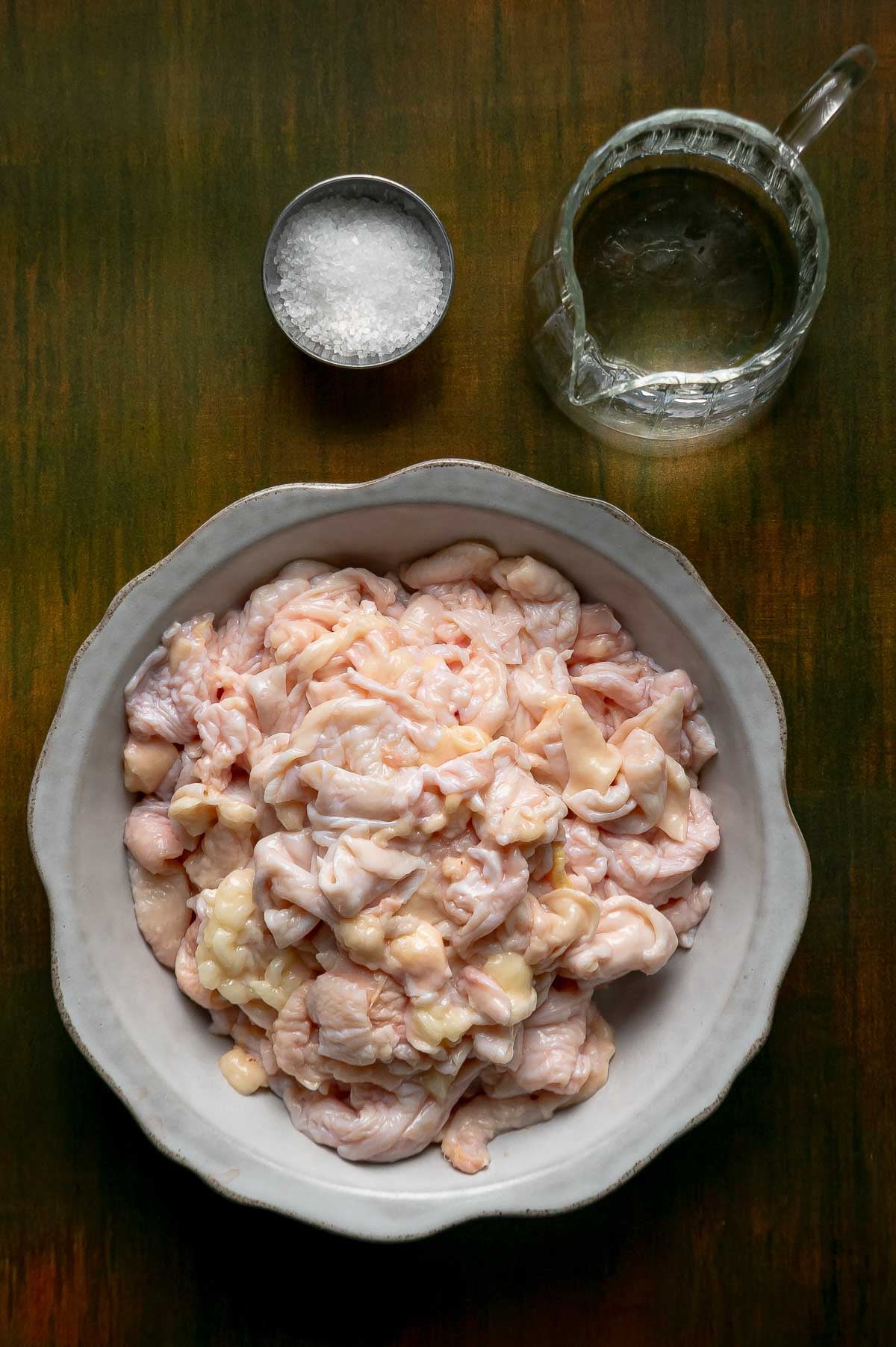 Ingredients for schmaltz and gribenes: a bowl of raw chicken skins cut into small pieces, a dish of kosher salt and a small glass pitcher of water.