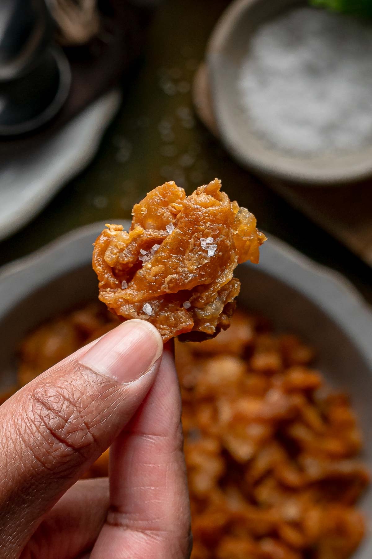 A hand holding up one piece of salted gribenes, crispy fried chicken skin, over the bowl.