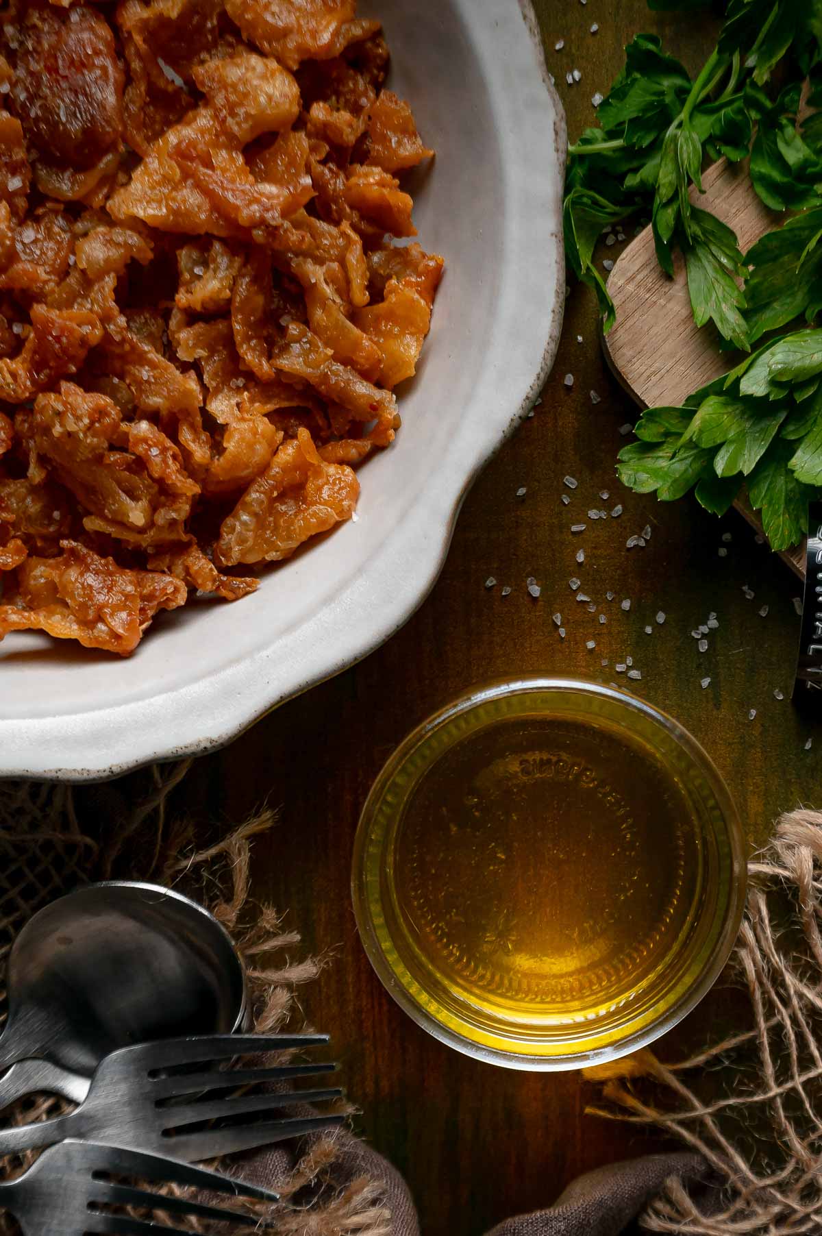 A bowl of gribenes beside a small glass bowl of liquid schmaltz, fresh parsley and forks on a dark table.