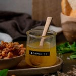 A labeled jar of homemade schmaltz with a wooden spoon beside a bowl of gribenes on a wooden tray.