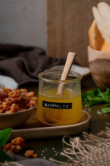 A labeled jar of homemade schmaltz with a wooden spoon beside a bowl of gribenes on a wooden tray.