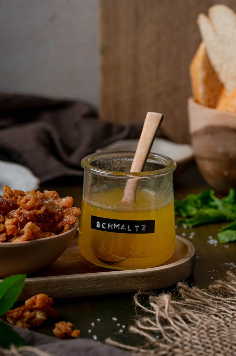 A labeled jar of homemade schmaltz with a wooden spoon beside a bowl of gribenes on a wooden tray.