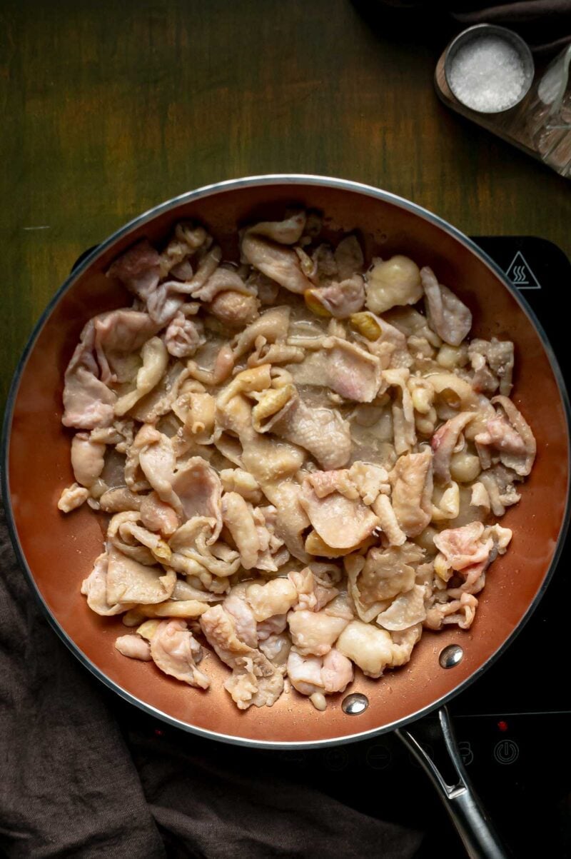Raw chicken skins spread in an even layer in a skillet at the start of rendering schmaltz.