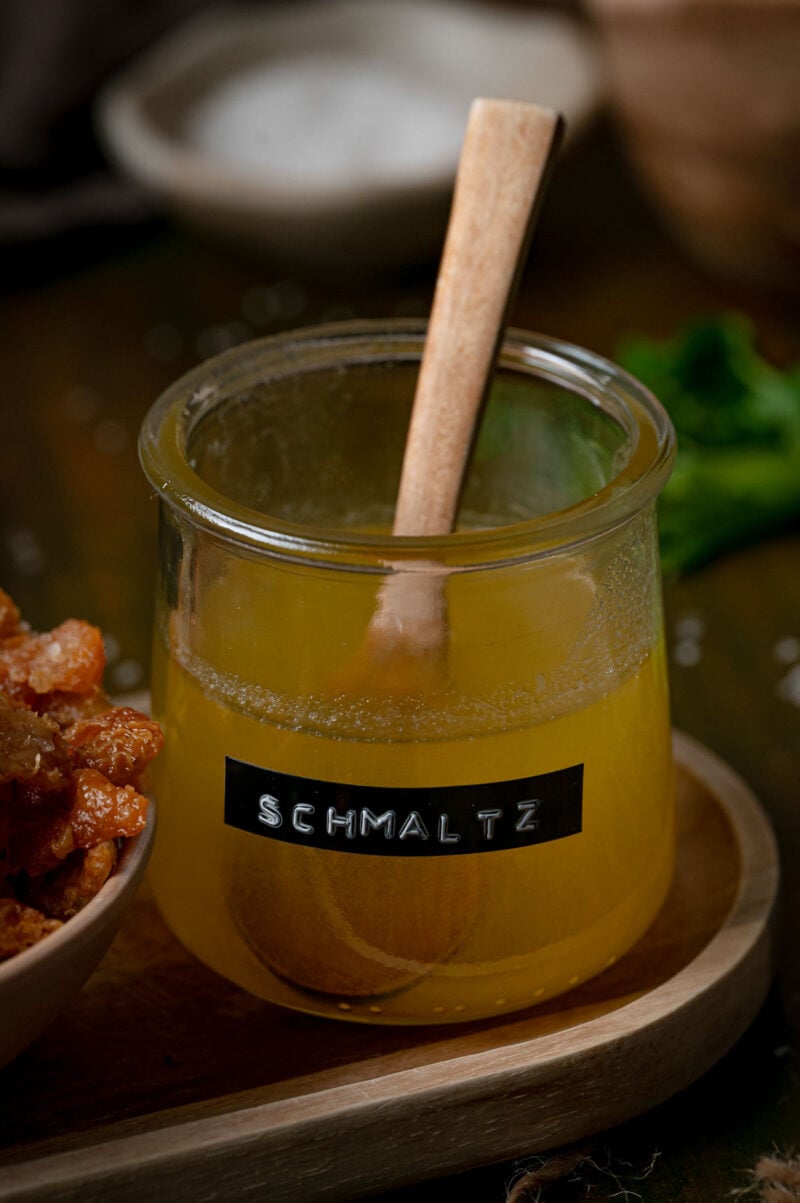 A glass jar of yellow schmaltz with a wooden spoon and a label reading “SCHMALTZ.”