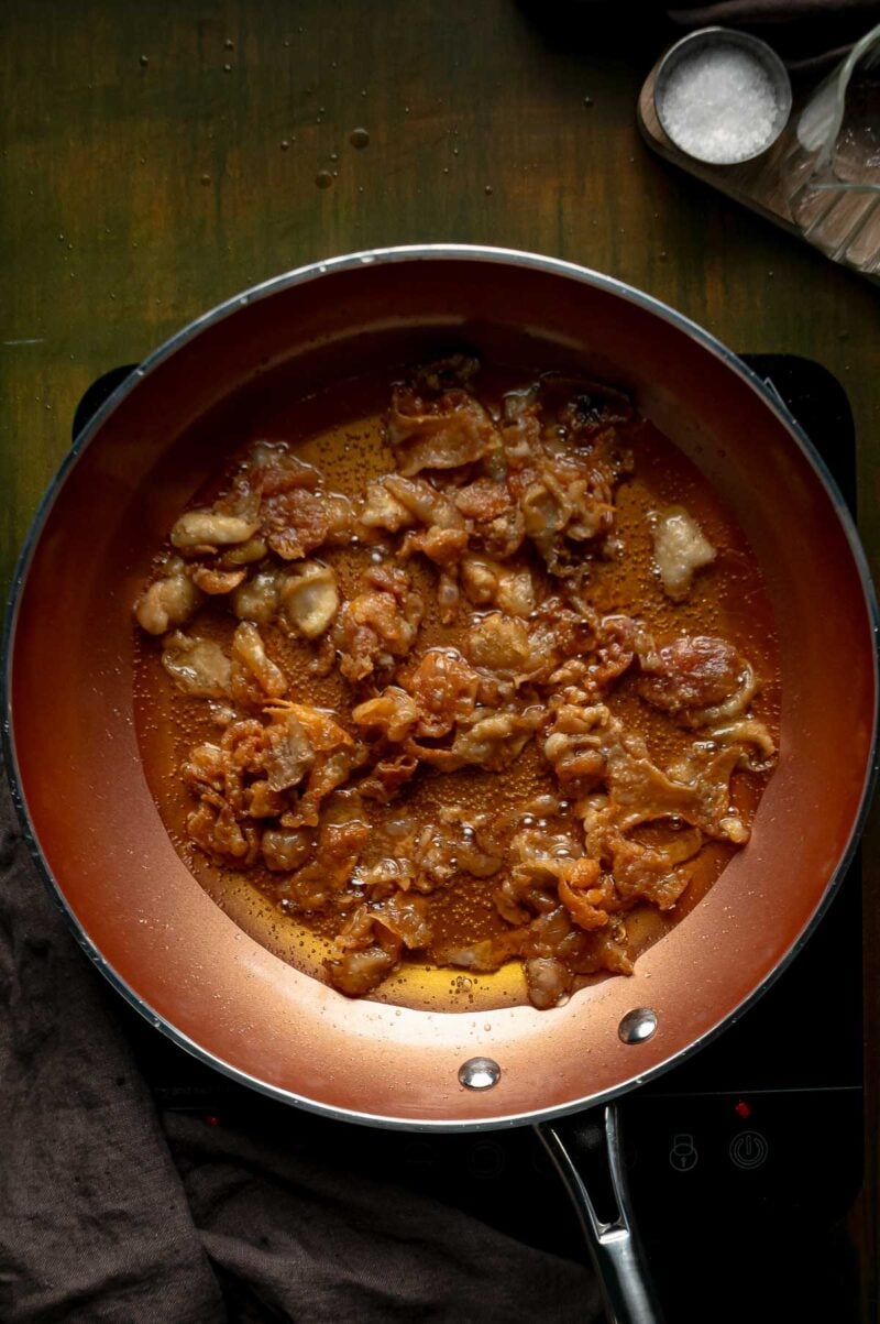 Chicken skins curling and browning in a skillet of rendered schmaltz, halfway to crispy gribenes.