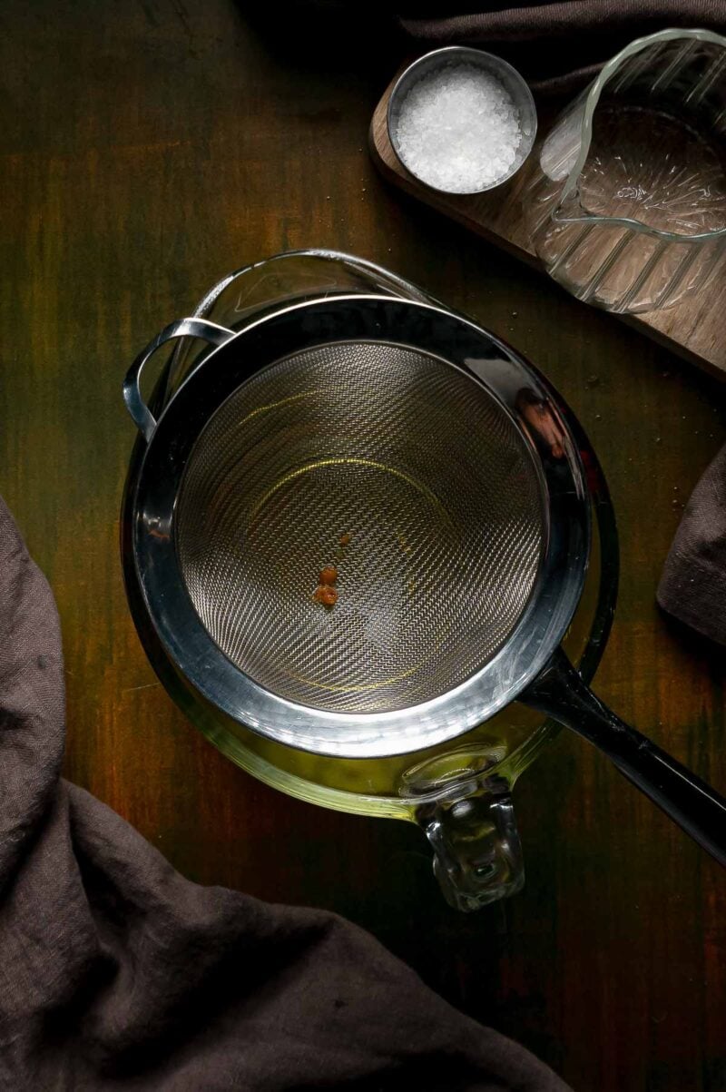 A fine mesh strainer set over a glass measuring cup of strained schmaltz, with one browned bit caught in the mesh.