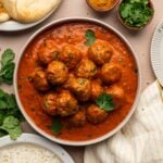 A bowl of creamy Indian lamb meatball curry with naan, basmati rice, lemon wedges, curry powder and fresh cilantro around it.