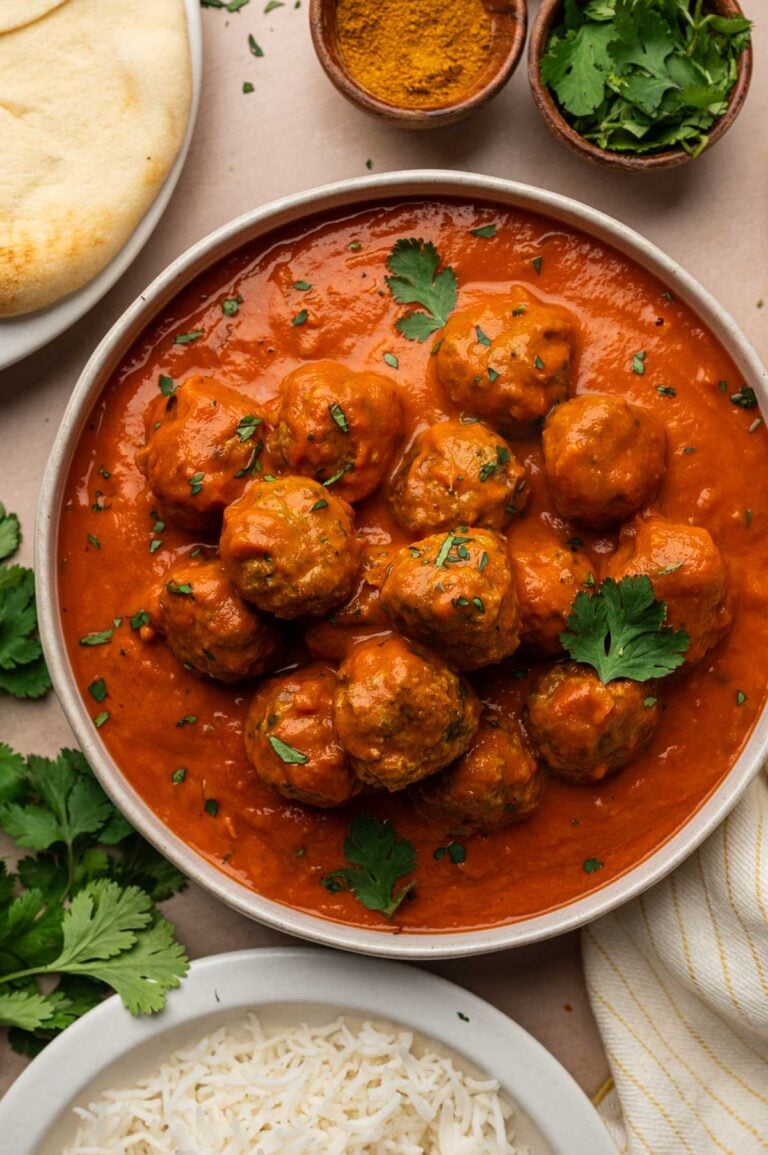 Overhead of a bowl of lamb meatball curry topped with cilantro leaves, with naan, basmati rice, curry powder and fresh cilantro.