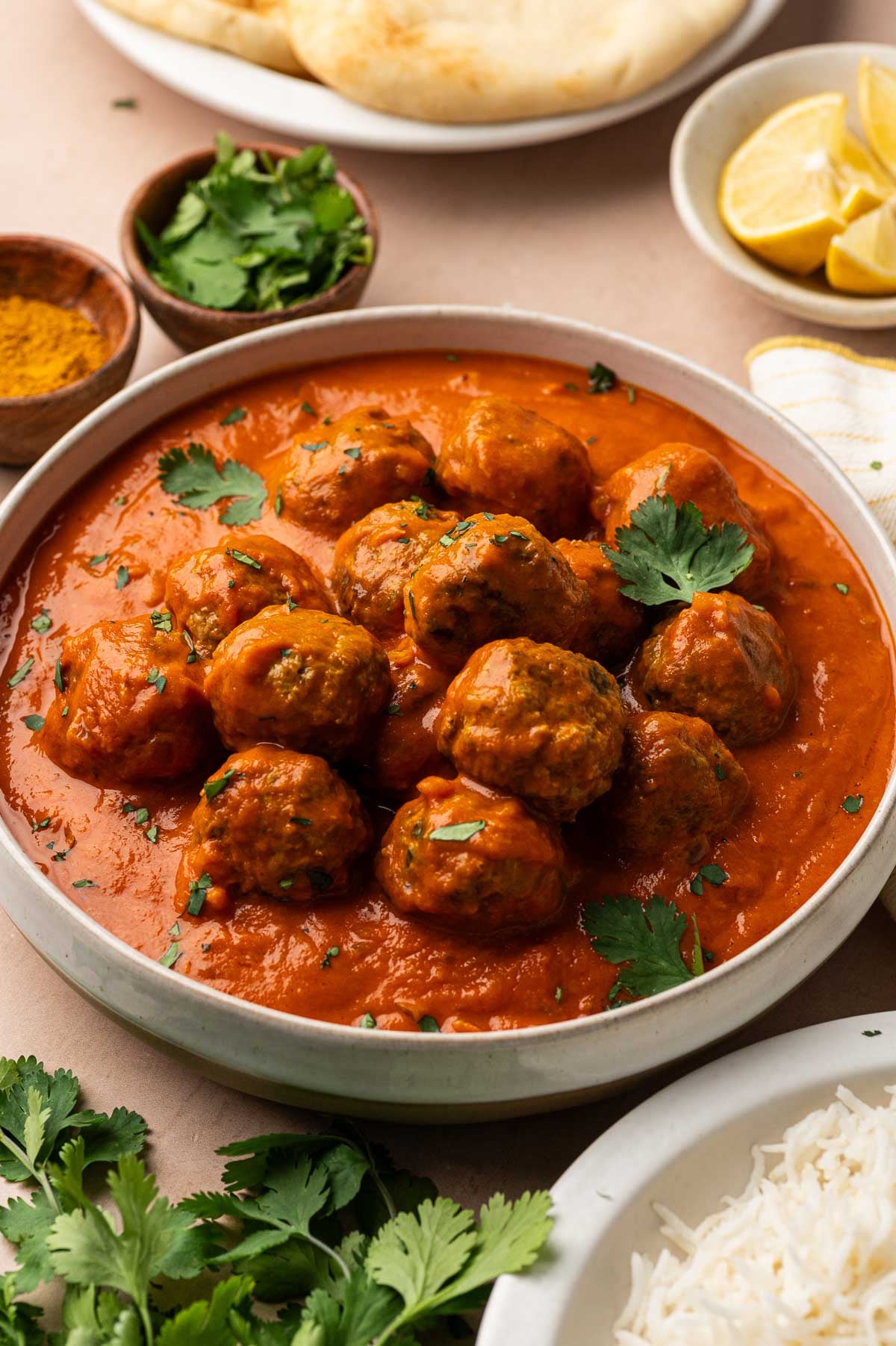 A bowl of lamb meatball curry with naan, lemon wedges, curry powder, cilantro and basmati rice on the side.