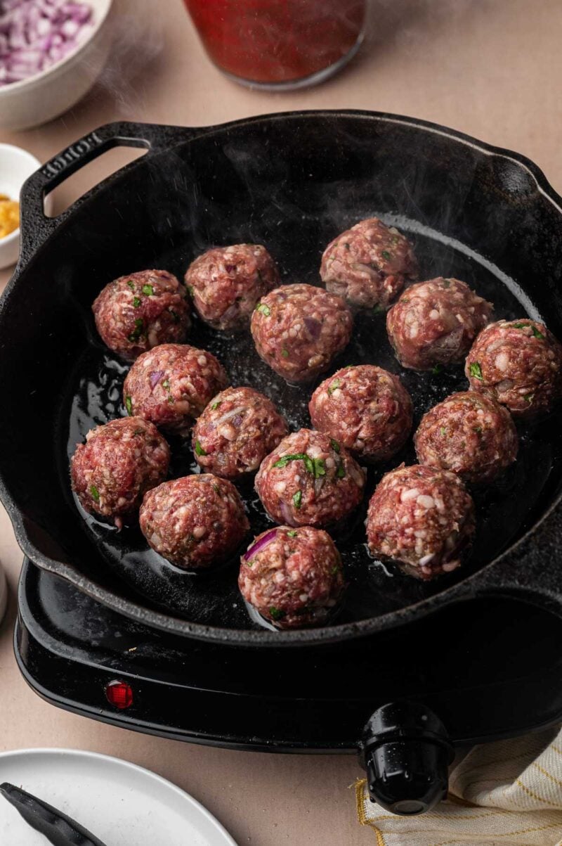 Raw lamb meatballs going into a hot cast iron skillet to brown.