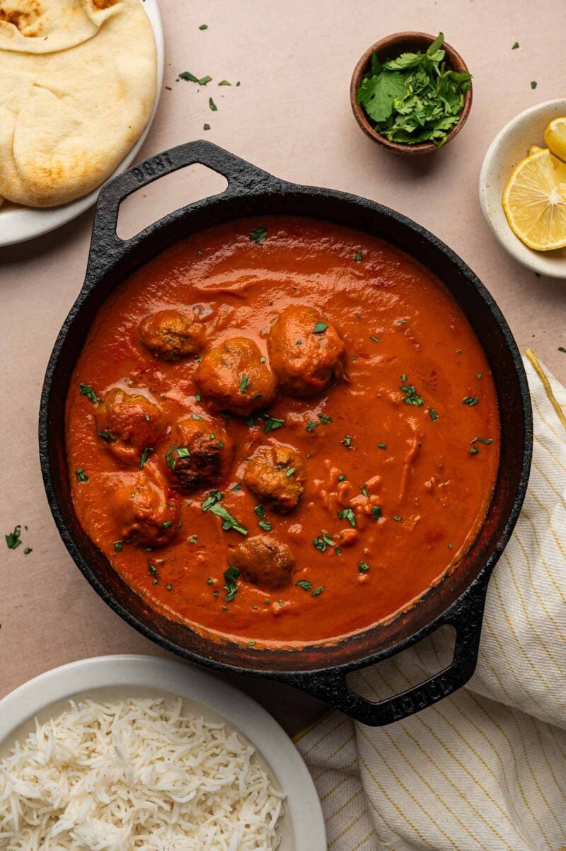 A cast iron skillet of lamb meatball curry in creamy tomato sauce with cilantro, naan, lemon wedges and a bowl of basmati rice.