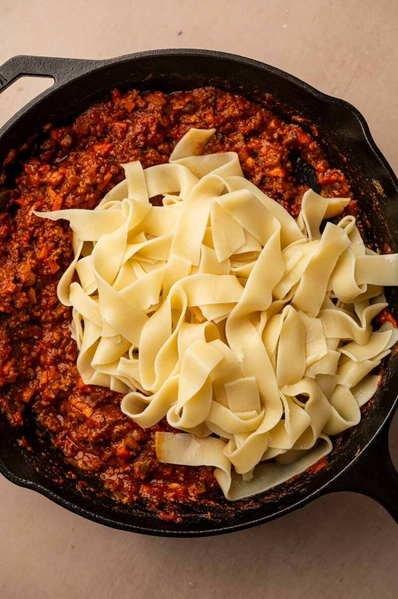 Drained pappardelle piled on top of the thick lamb bolognese in the cast iron skillet, ready to toss.