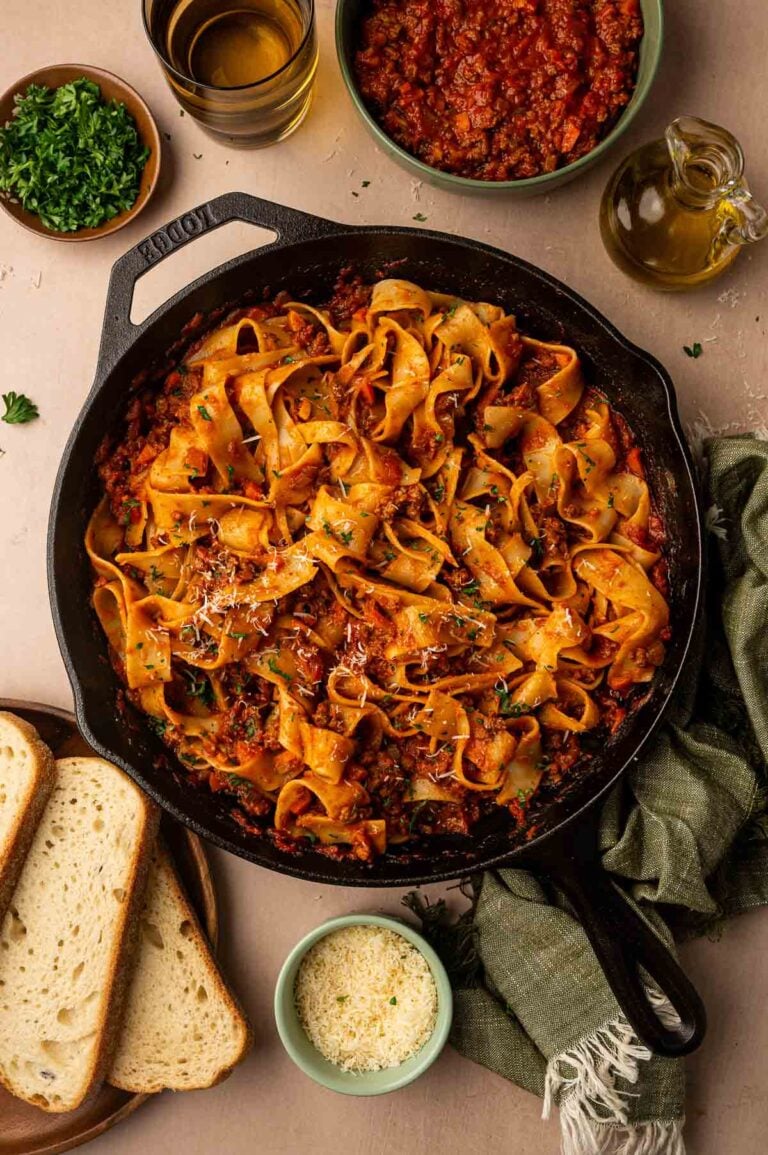 Lamb bolognese tossed with pappardelle in a cast iron skillet, surrounded by sliced bread, chopped parsley, grated Parmesan and a bowl of extra sauce.