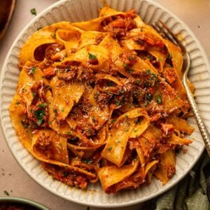 A bowl of lamb bolognese with pappardelle and a fork, next to the cast iron skillet and a bowl of extra lamb sauce.