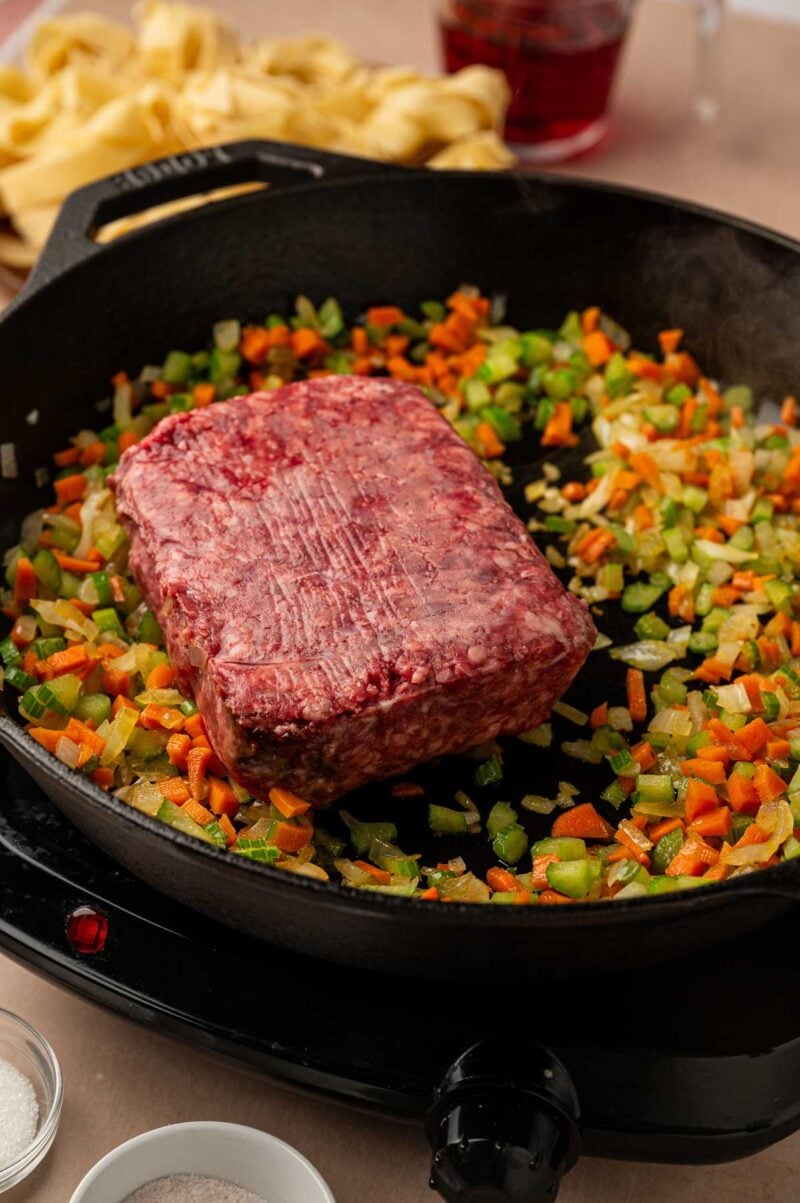A block of ground lamb set on top of the softened onion, carrots and celery in the cast iron skillet, ready to brown.