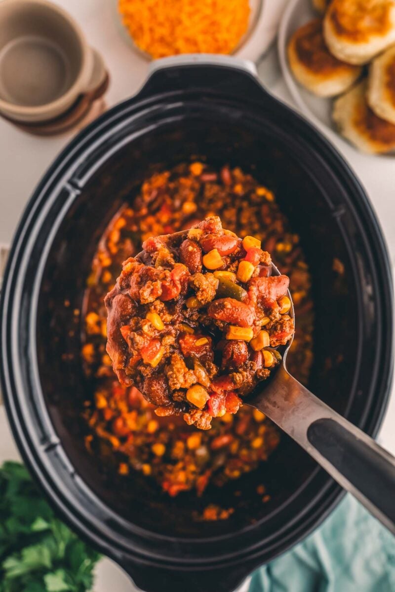 A ladle lifting thick slow cooker turkey chili with kidney beans and corn from the crock, with biscuits and shredded cheddar nearby.