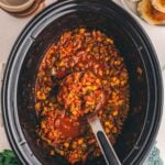 Finished slow cooker turkey chili in the crock with a ladle, set out with biscuits, bowls and shredded cheddar.