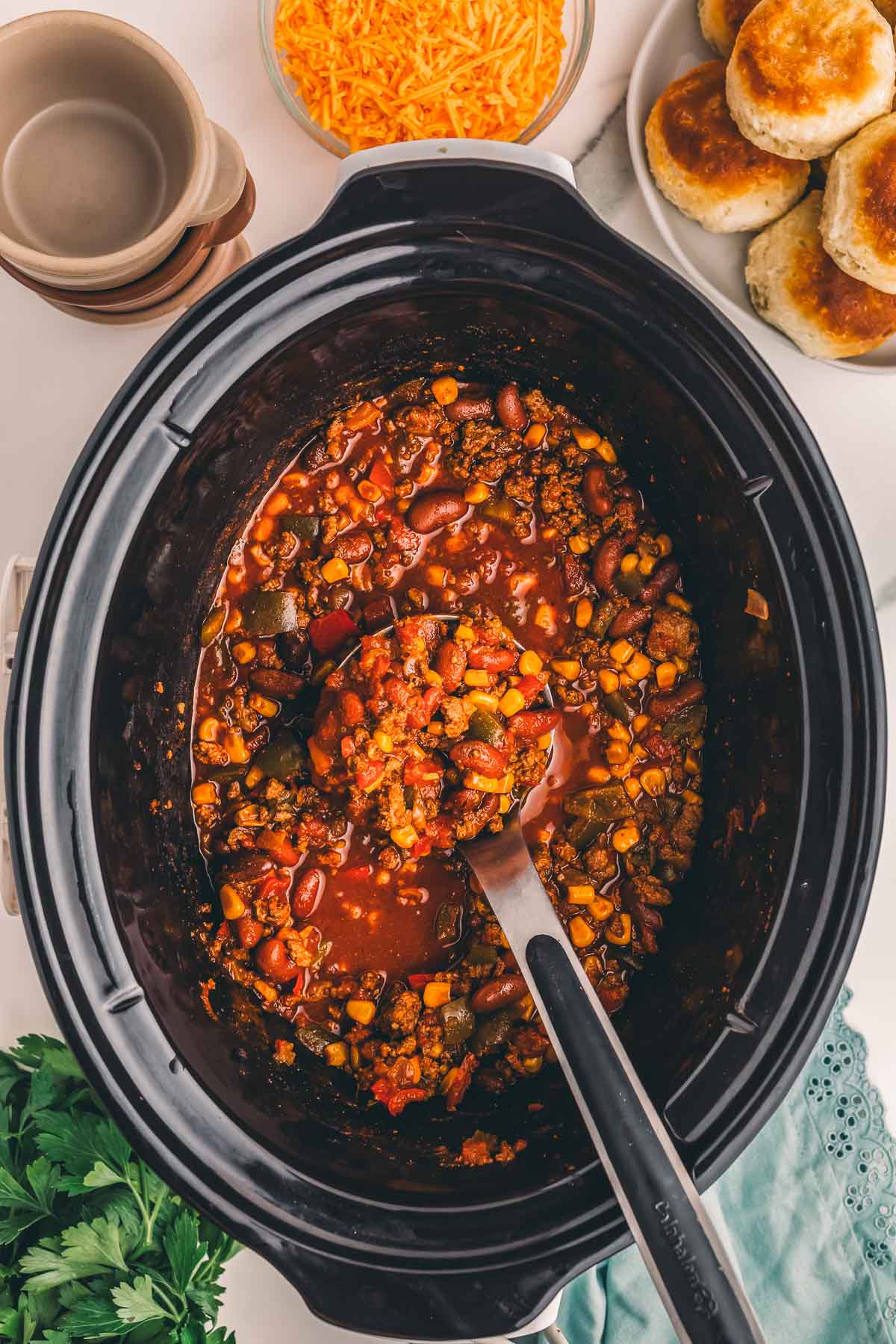Finished slow cooker turkey chili in the crock with a ladle, set out with biscuits, bowls and shredded cheddar.