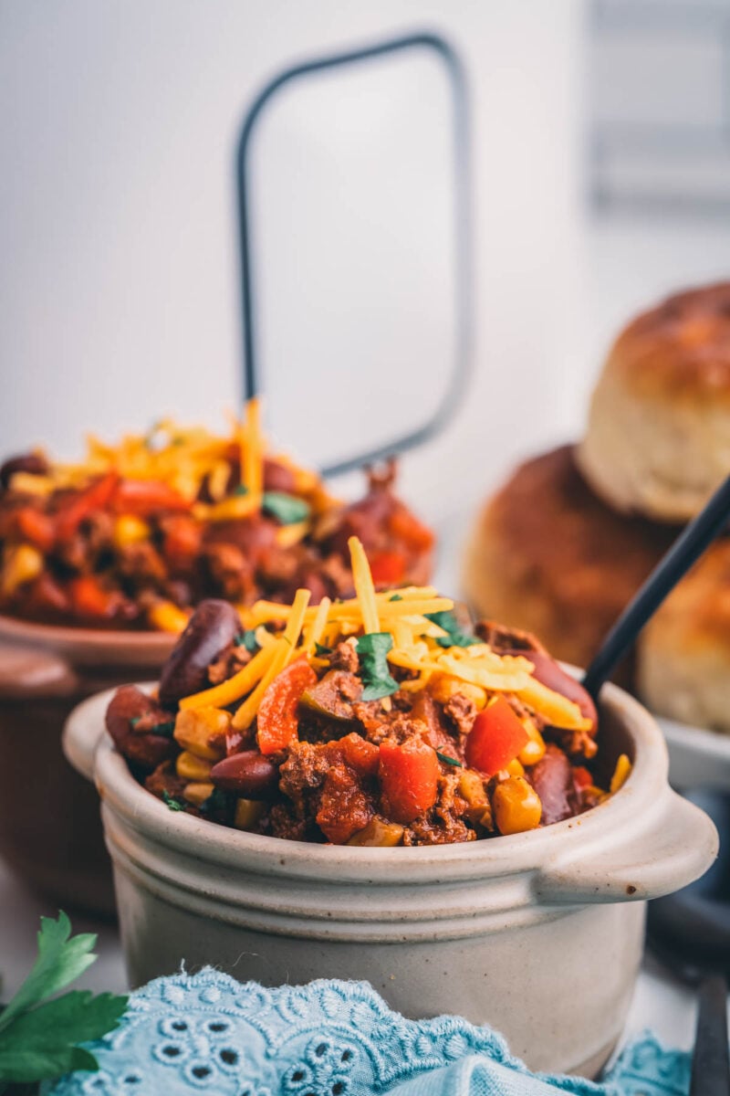 Close up of a bowl of slow cooker turkey chili with kidney beans and shredded cheddar, biscuits in the background.